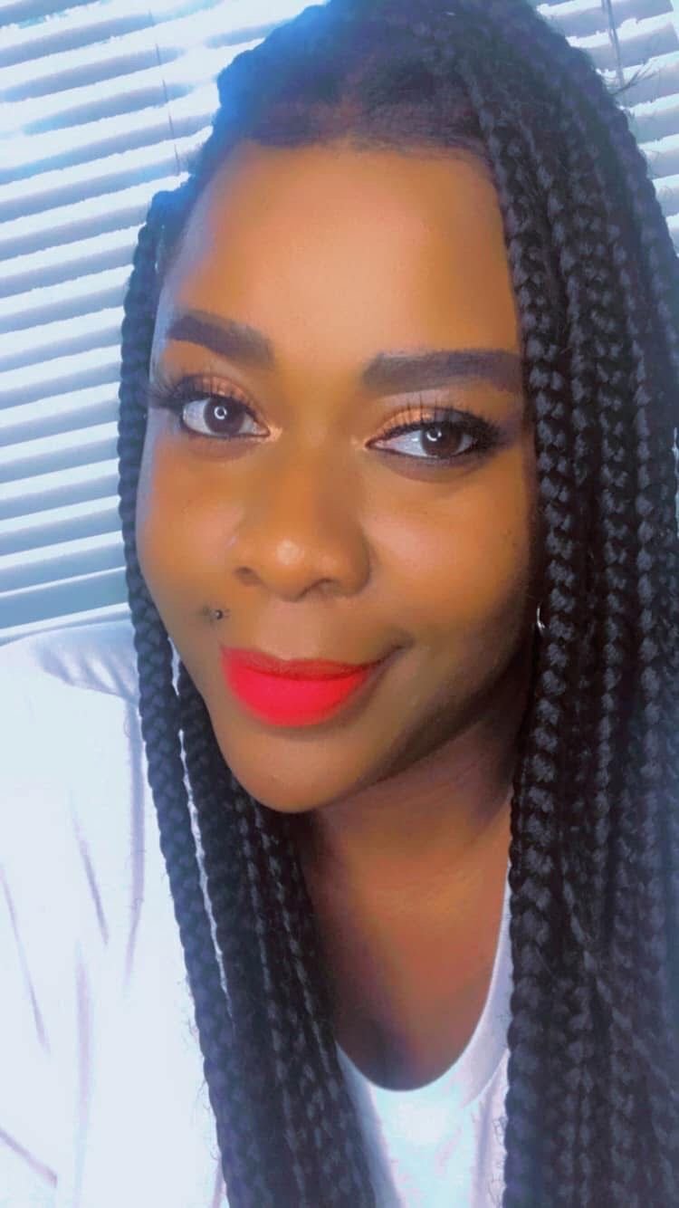 Woman with braided black hair, red lipstick, and orange eyeshadow smiling at camera against a white venetian blind background