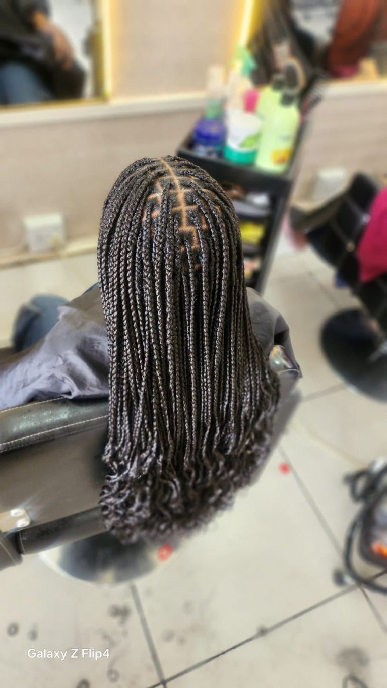 Back view of a person with long braided hair in a salon chair with mirrors and colorful items visible in the background